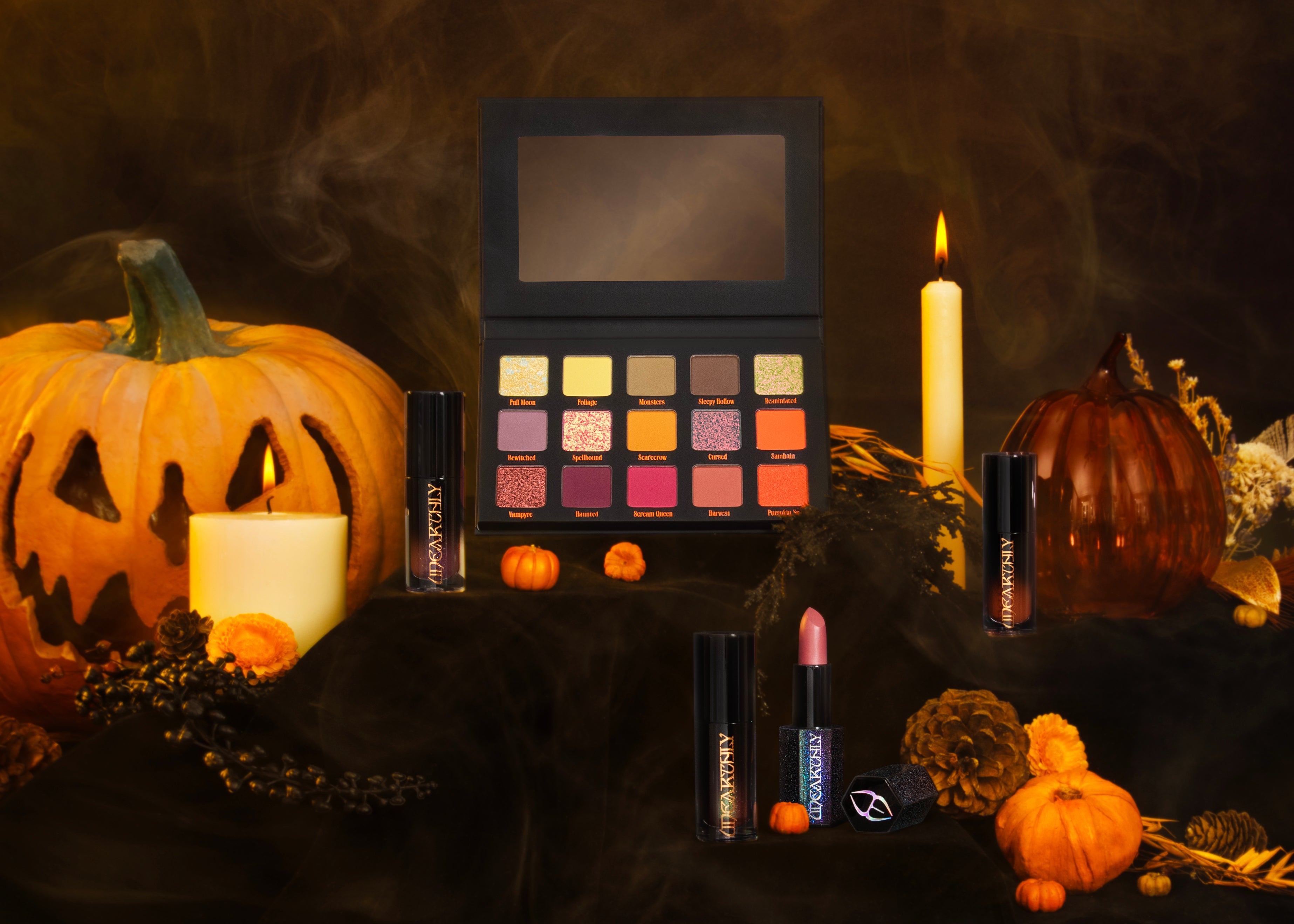 A Halloween-themed beauty display featuring an open makeup palette with 16 shades, two lipsticks, and two tubes of eyeshadow, arranged with pumpkins, candles, and pinecones on a dark draped surface.