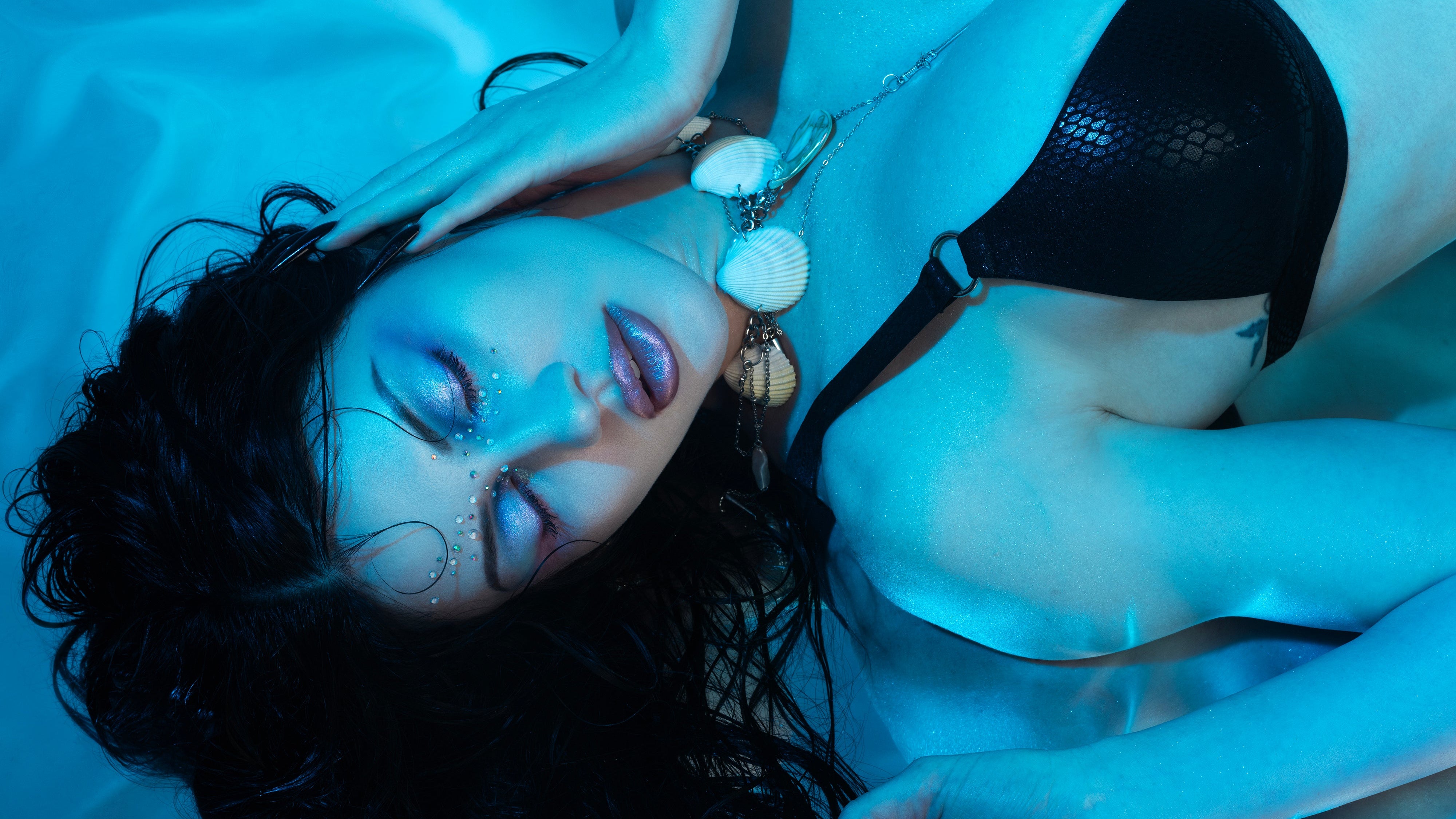 A woman lying on her back under blue-toned lighting, wearing a black mesh bikini top and a shell pendant necklace.